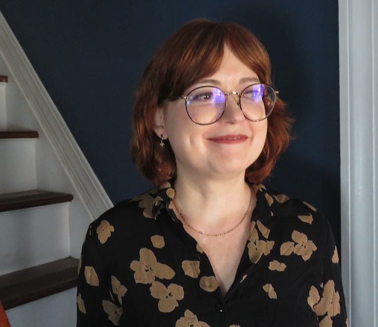 Image of Maud Newton, a bespectacled white woman in a black blouse with brown flowers, standing in front of stairs. Photograph by Maximus Clarke.