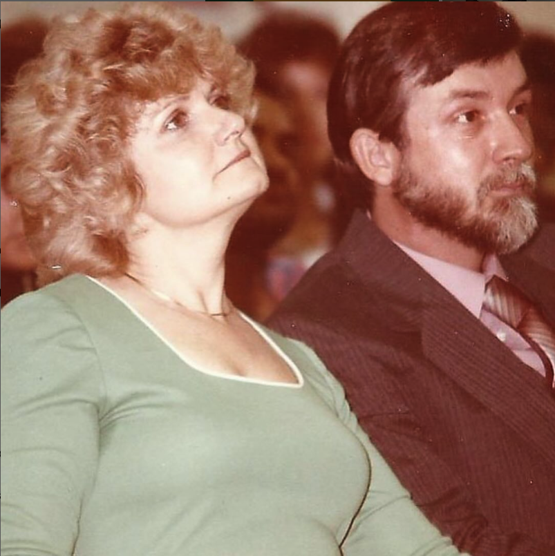 Image shows a white woman in a pale green dress and man in a suit seated and looking on with approval.